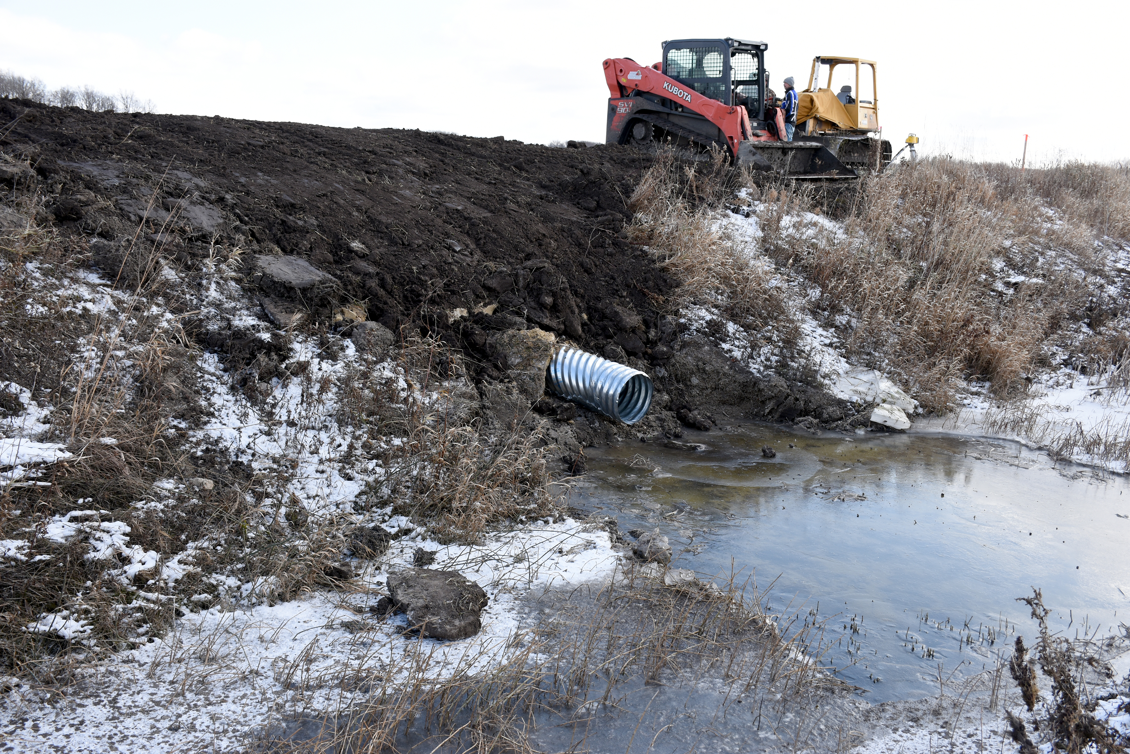 A metal pipe extends from a ditch bank to a ditch with open water. Construction equipment sits at the edge of a field.