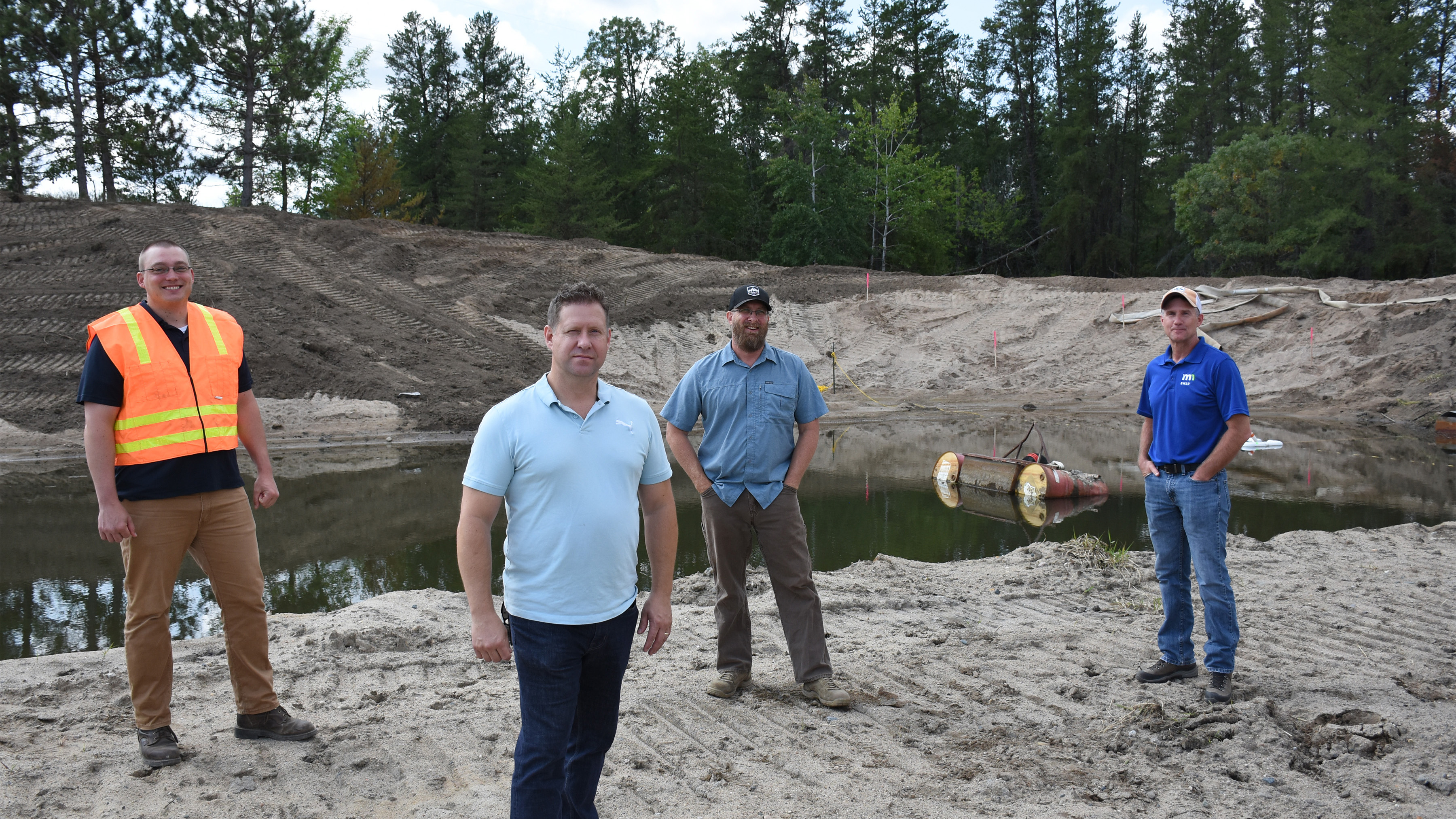 Four men stand in front of a pond filled with water