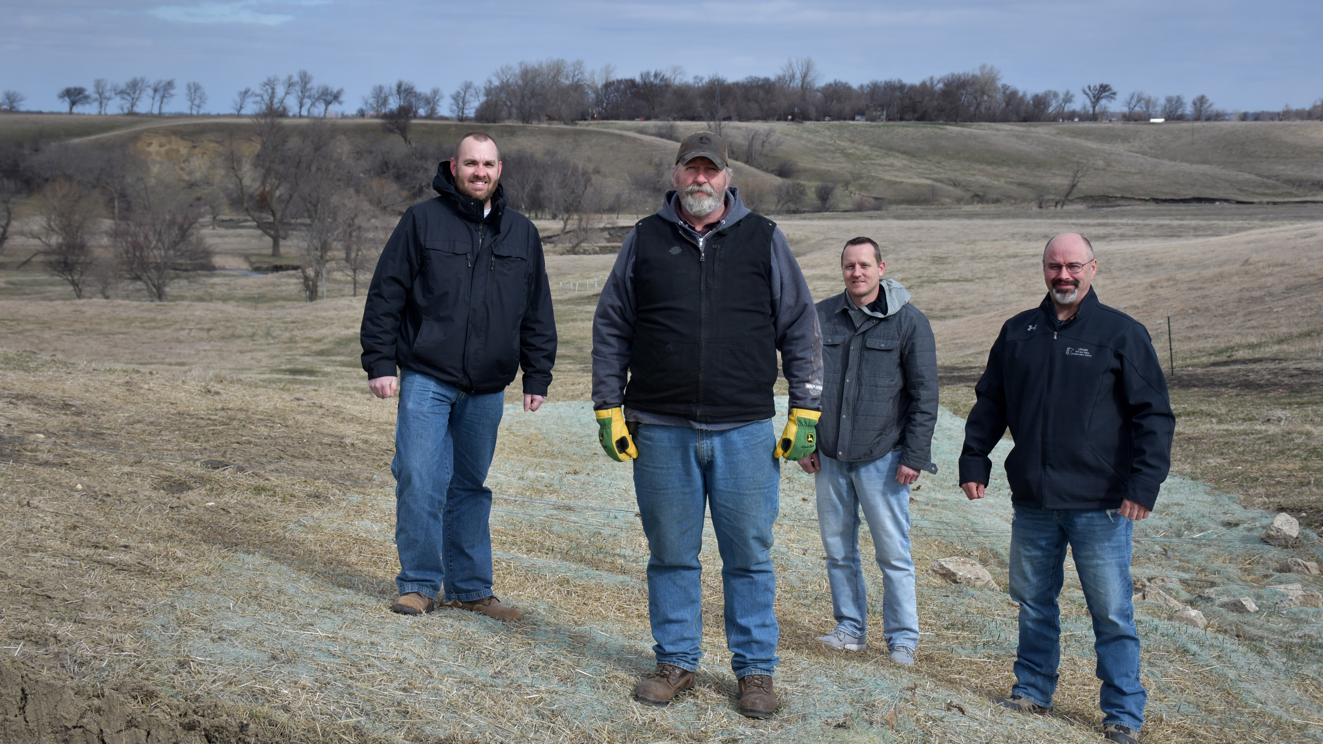Four men stand in front of a fence with hills and trees in the background and erosion control fabric in the foreground