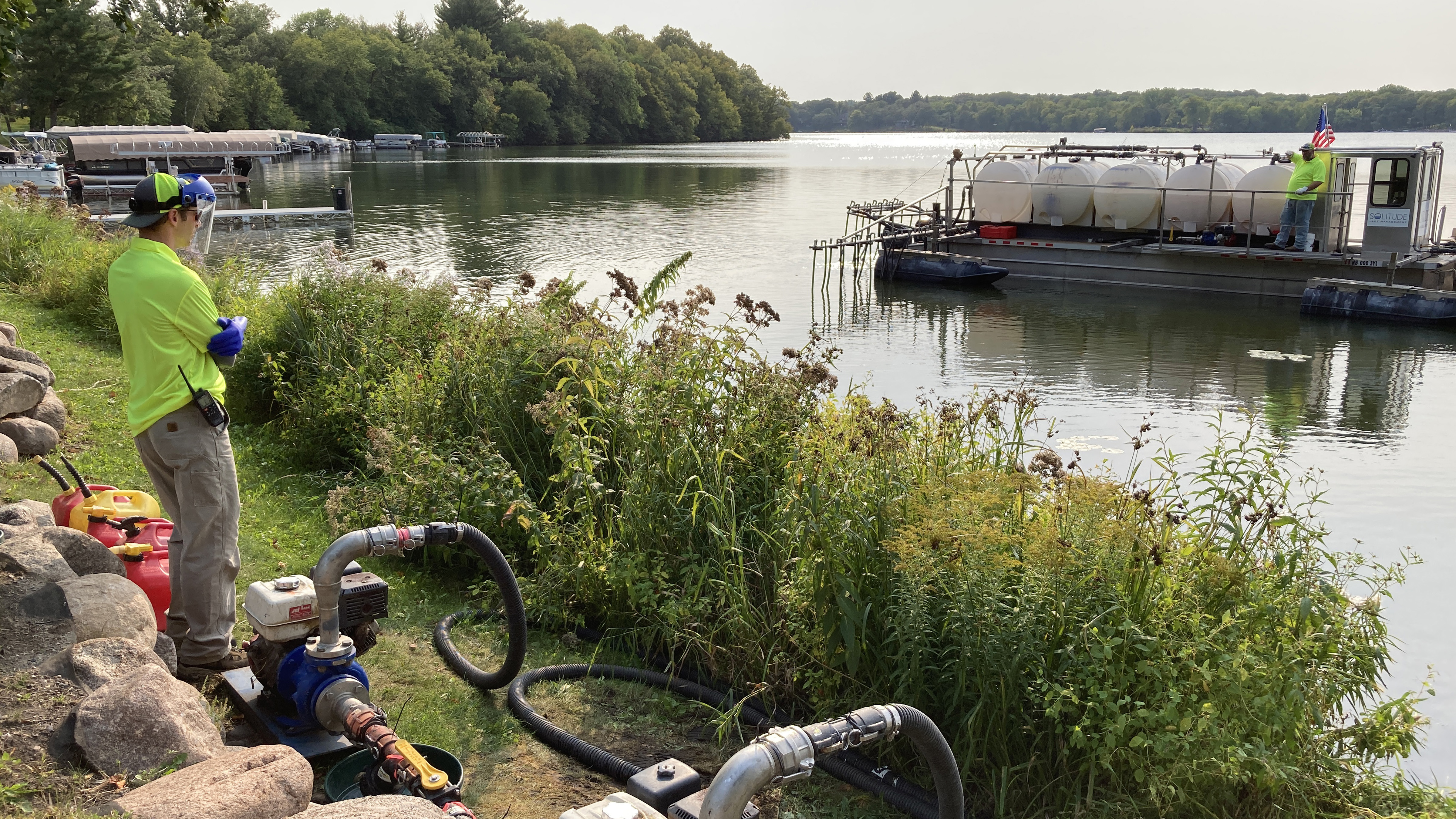 Pumps and hoses are on the lakeshore where a man watches a barge with white plastic tanks being filled
