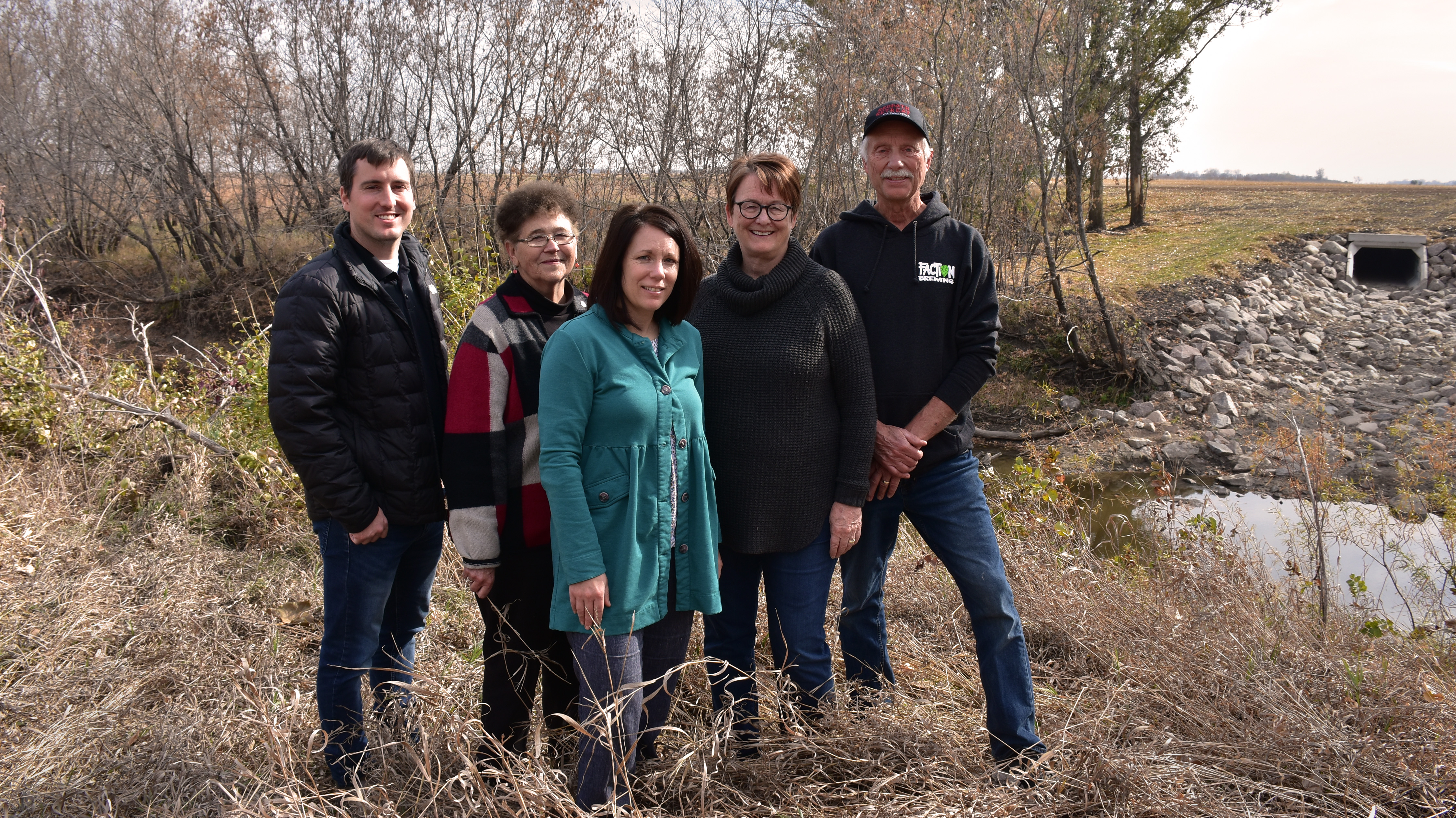 Five people stand in a group with a creek and a large square culvert in the background
