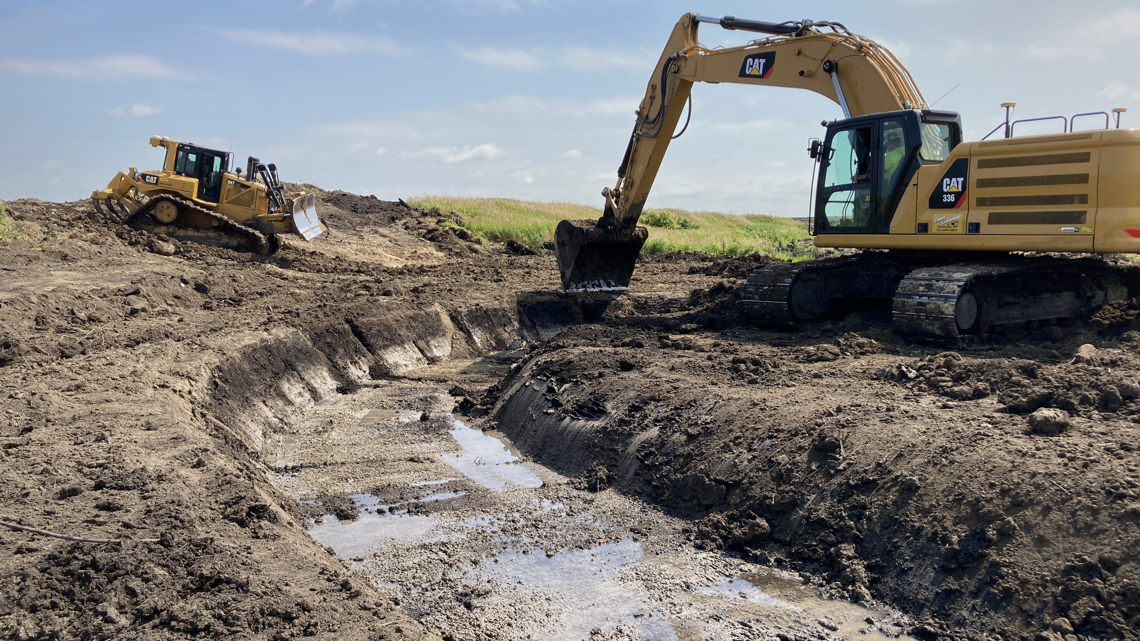 A backhoe excavates a stream channel. A bulldozer is in the background.