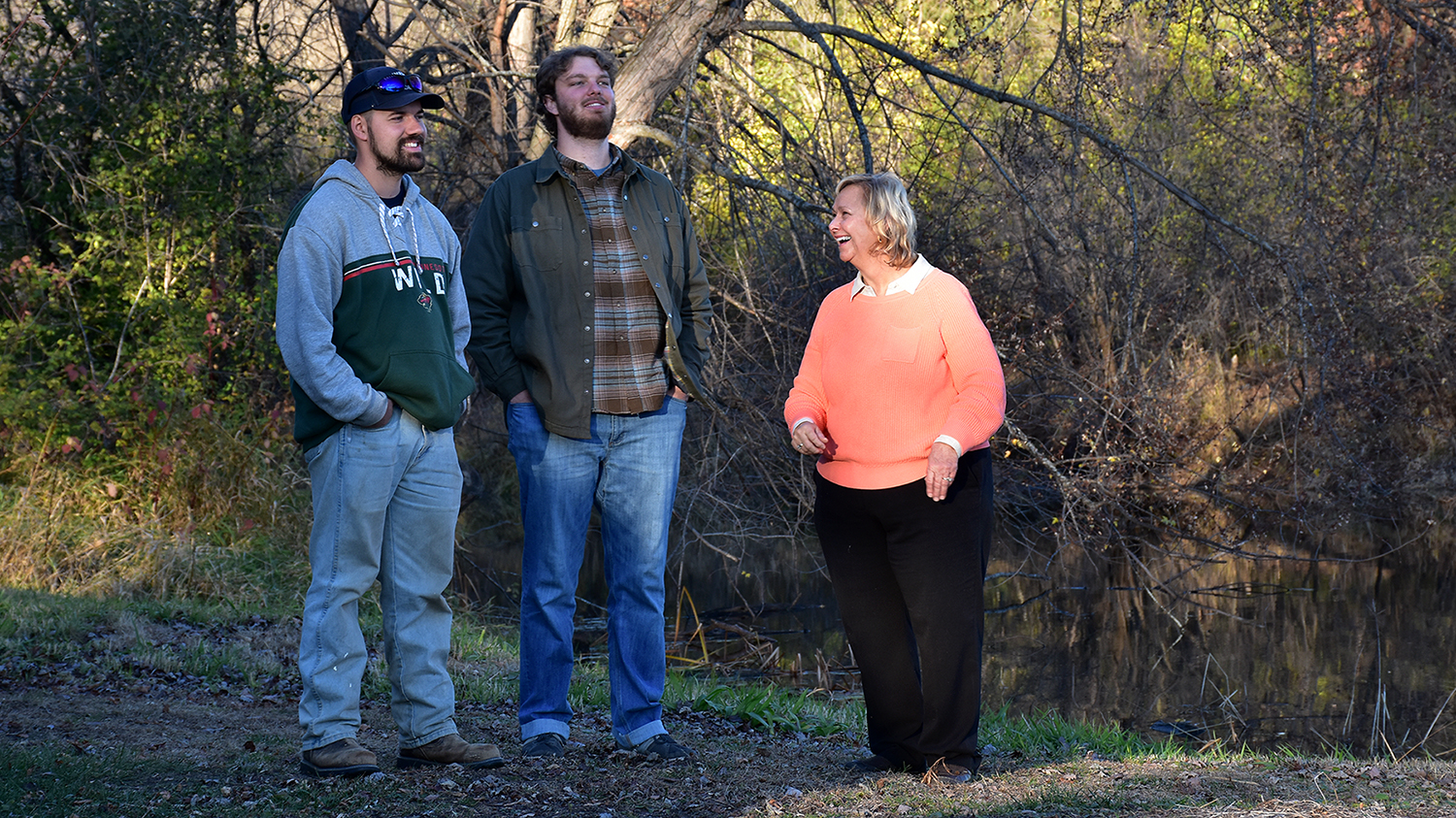 three people stand by a river with trees in the background