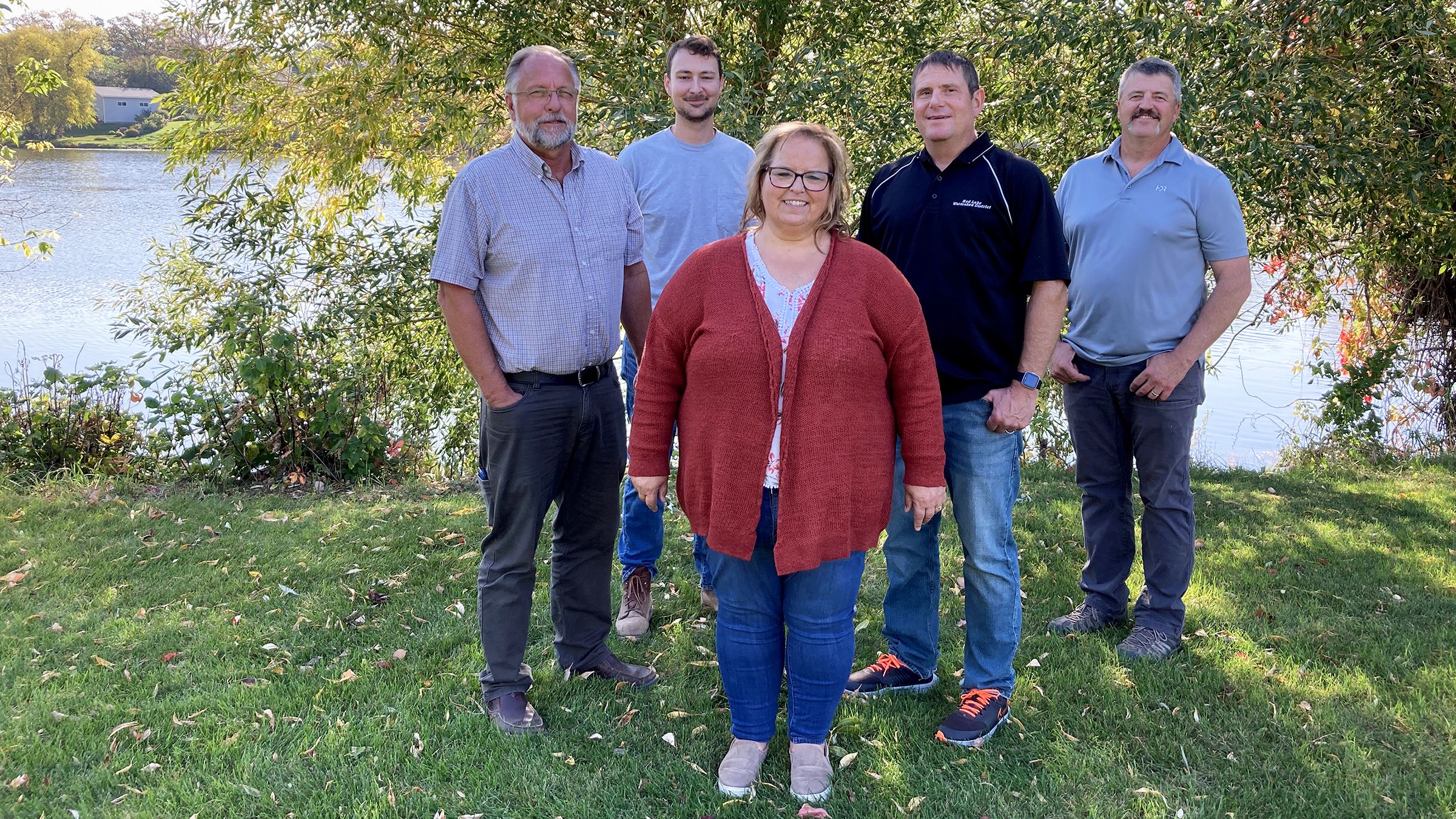 Five people stand on the grassy bank of a river with trees in the background