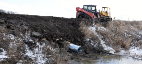 A metal pipe extends from a ditch bank to a ditch with open water. Construction equipment sits at the edge of a field.