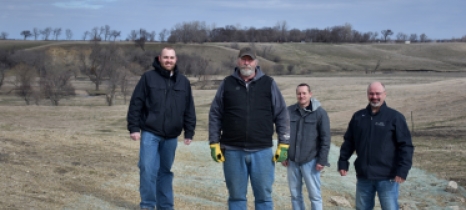 Four men stand in front of a fence with hills and trees in the background and erosion control fabric in the foreground