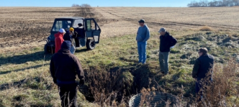 A group of people standing at the edge of a field viewing a hole in the ground.