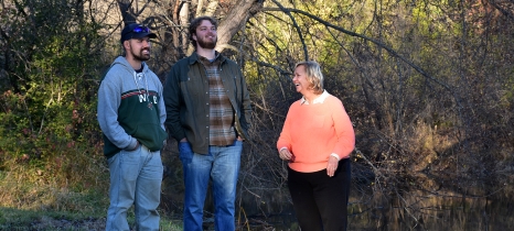 three people stand by a river with trees in the background