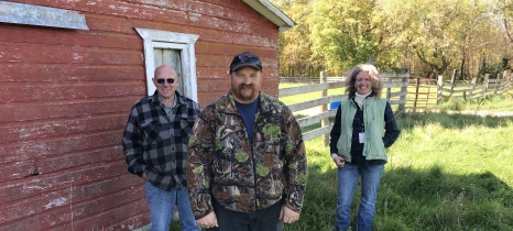 three people stand along the side of a red barn. a wooden fence is in the background