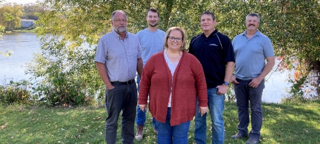 Five people stand on the grassy bank of a river with trees in the background