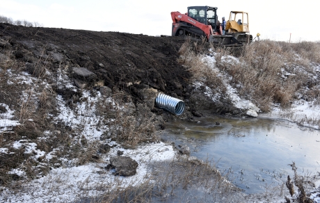 A metal pipe extends from a ditch bank to a ditch with open water. Construction equipment sits at the edge of a field.