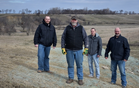 Four men stand in front of a fence with hills and trees in the background and erosion control fabric in the foreground