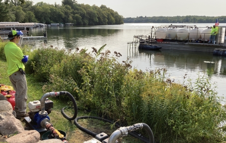Pumps and hoses are on the lakeshore where a man watches a barge with white plastic tanks being filled