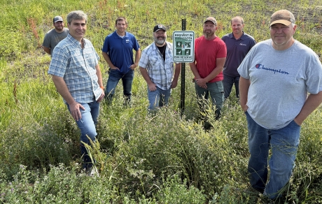 Seven people stand in tall plants around a Reinvest in Minnesota Reserve sign.